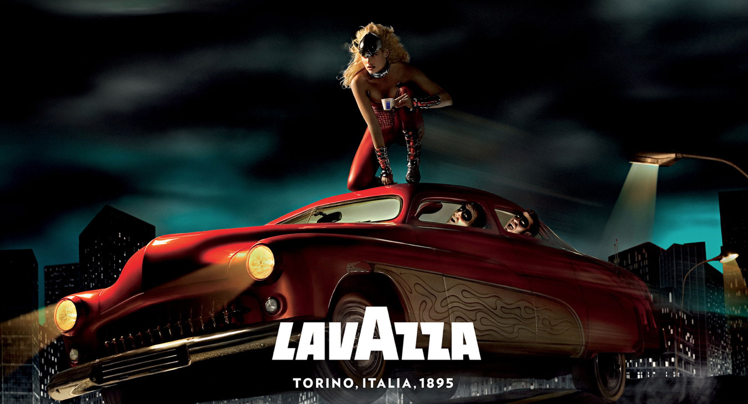 Lavazza advertising image — a model in red on the roof of a red vintage car at night, city skyline behind, with the Lavazza wordmark and “Torino, Italia, 1895”